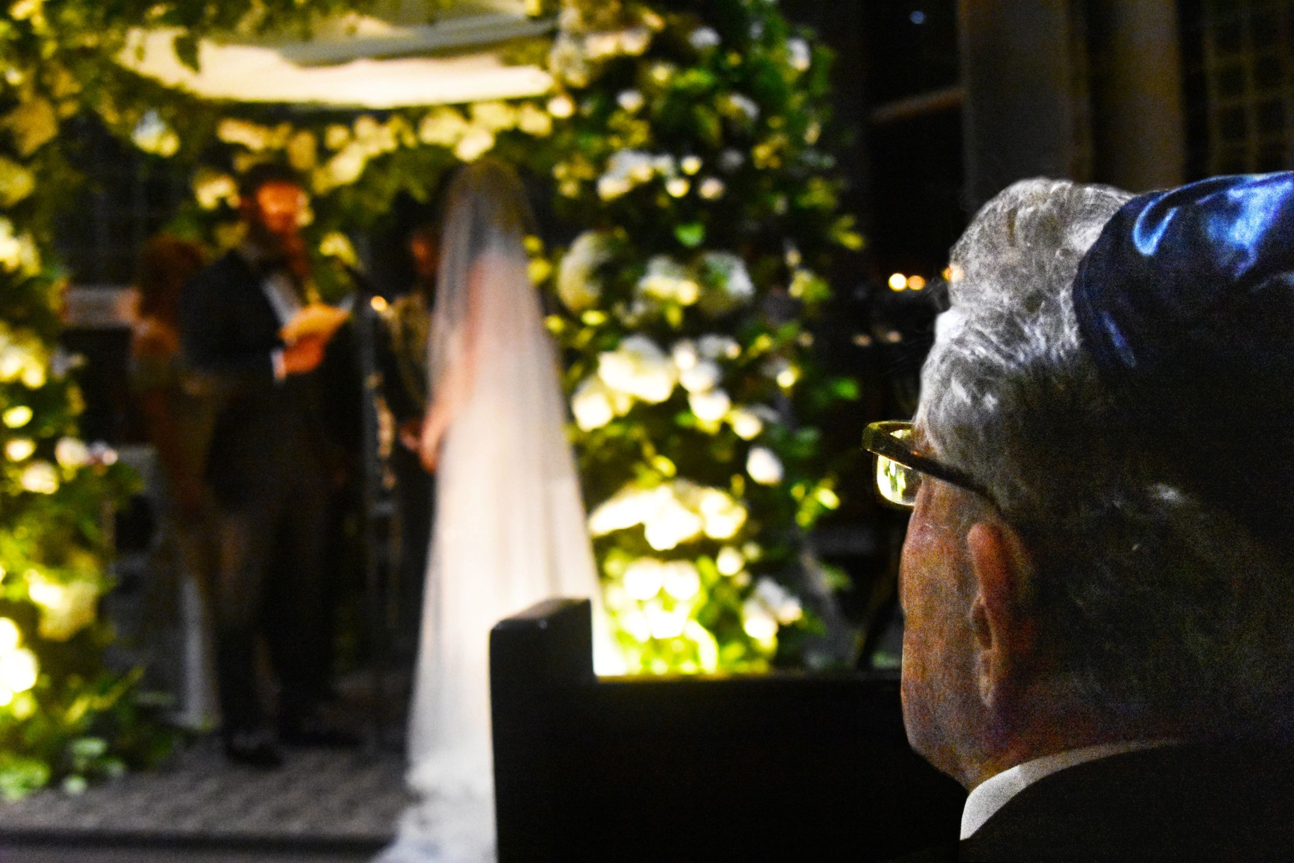 An elderly man watches a wedding ceremony with a bride and groom at a floral altar.