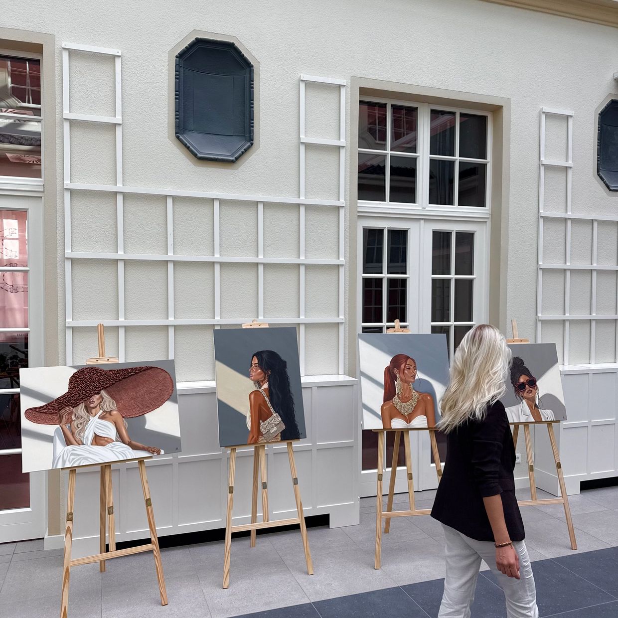 A woman with blonde hair observes four paintings of stylish women on easels indoors.