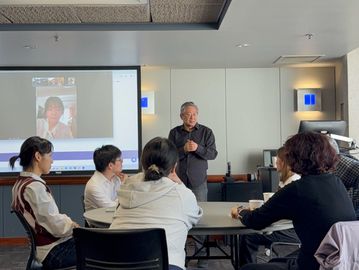 A man speaks to a group seated around a table with a video call projected behind.