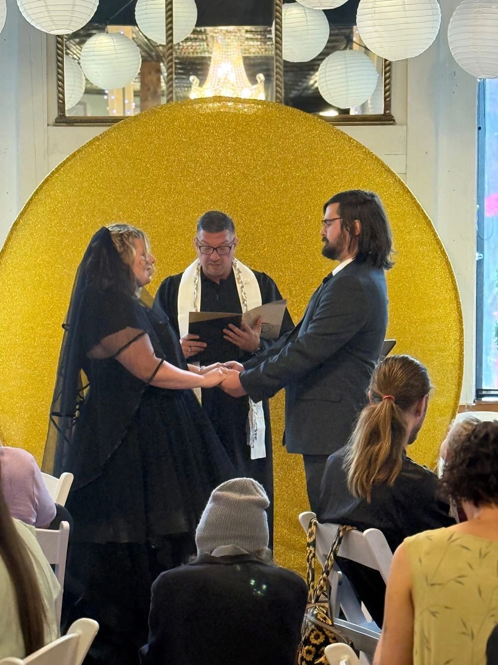 Couple in black attire exchanging vows with an officiant in front of a golden backdrop.