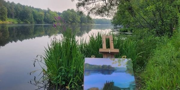 A painting on an easel captures a serene lakeside view surrounded by lush greenery.