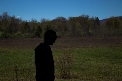 Silhouette of a person in a hat against a field and forest backdrop under a clear blue sky.