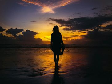 Woman walking along beach towards the sunset.