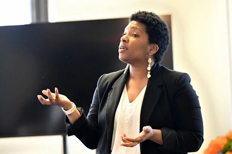 Whitney stands in a black suit jacket with her hands upturned. She is a Black woman with an afro.