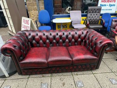 Worn red leather Chesterfield sofa displayed outdoors with various chairs in the background.