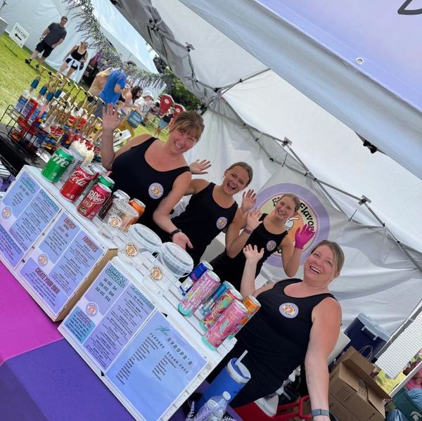 Four women cheerfully posing behind a colorful drink stand at an outdoor event.