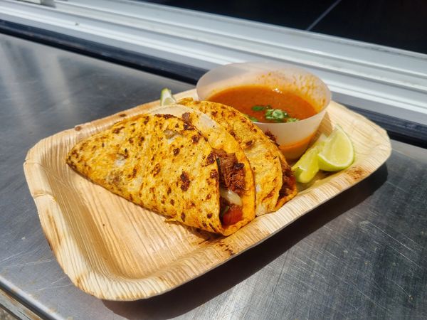 Three grilled tacos served with lime wedges and a cup of red salsa on a wooden tray.