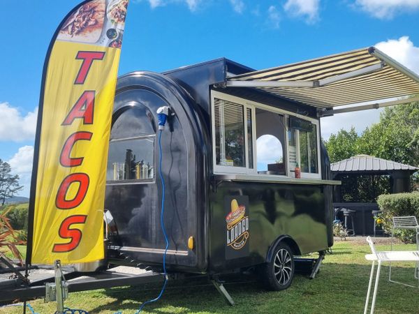 Black taco food truck with a yellow "TACOS" flag and striped awning outdoors.