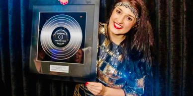 Woman proudly holding a framed gold record award, smiling at the camera.
