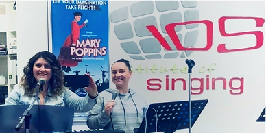 Two women smiling with microphones in a music studio with a keyboard and Mary Poppins poster.