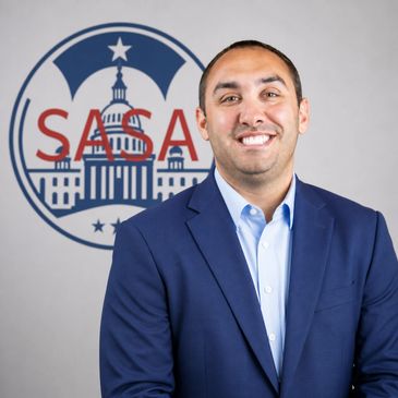 Man in a blue suit smiling in front of a SASA logo.