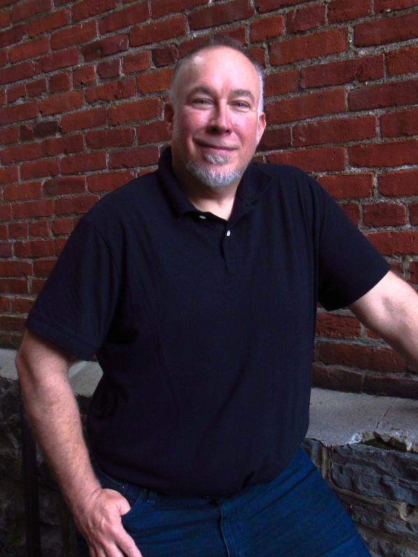 Smiling man in black polo shirt against brick wall.