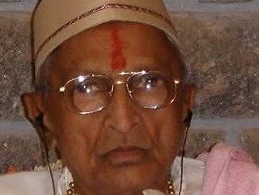Elderly man wearing traditional attire and glasses with a red tilak on his forehead.
