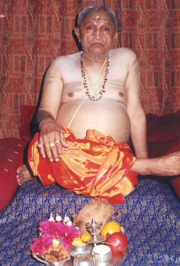 An elderly man in traditional attire seated with ritual items before him.