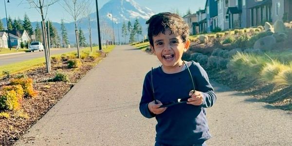 Smiling boy holding sunglasses on a sunny sidewalk.