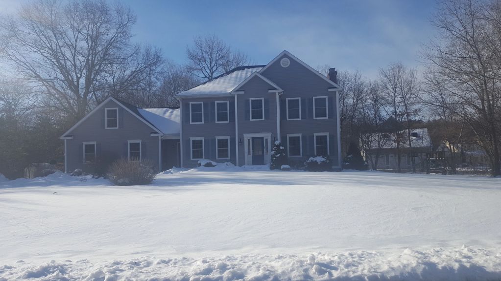 A gray two-story house with snow-covered yard under clear blue sky.