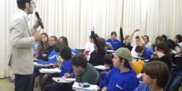A teacher speaking to a classroom of attentive students, some raising their hands.