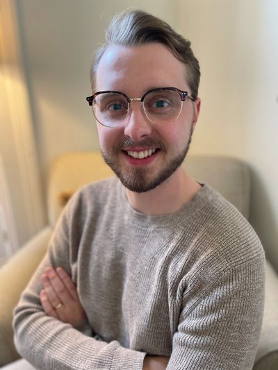 Smiling man wearing glasses and a beige sweater, sitting with arms crossed.