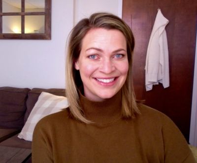 A woman smiling indoors wearing a brown turtleneck sweater.