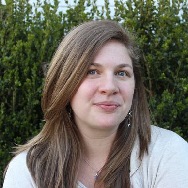Woman with light brown hair smiling gently outdoors near greenery.