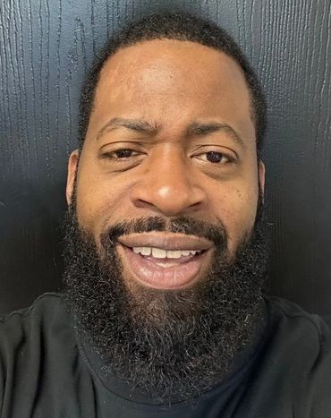 Smiling man with beard and curly hair in front of a black wall.