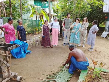 A group watches a man weaving palm leaves outdoors.