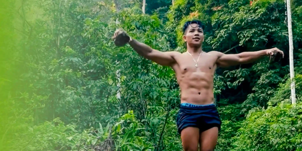 A shirtless man standing with arms open in a green forest.