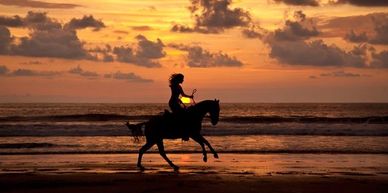 Person riding horse on beach at sunset.