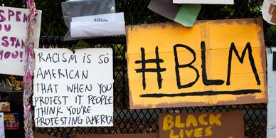 Picture of Protest signs yellow sign with Black Lettering #BLM, sign underneath is partially obscure