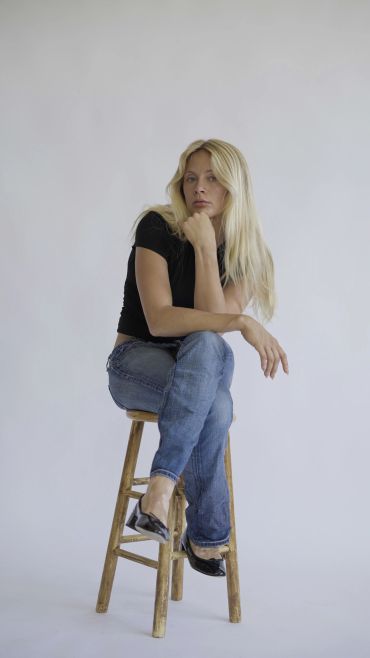 Woman sitting on a wooden stool with a thoughtful expression.