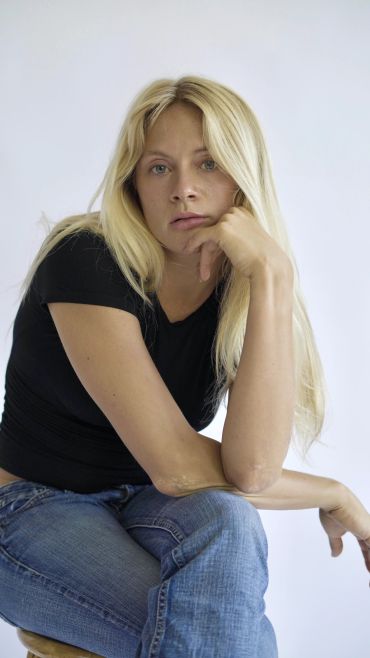 Blonde woman in black shirt and jeans sitting thoughtfully.