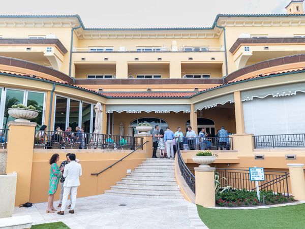 People socializing on the terrace of a large, elegant building with stairs and planters.
