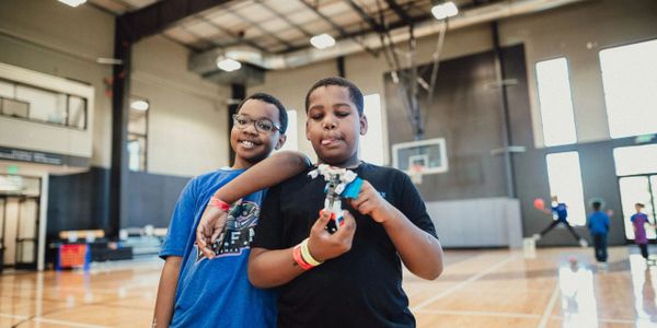 Two smiling boys at Love Me For Me after-school program in Bremerton, WA, enjoying a hands-on STEM activity with LEGO robotics inside a community gym