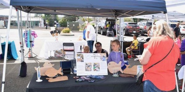 Outdoor craft booth with wooden fish cutouts and informational display under a canopy.