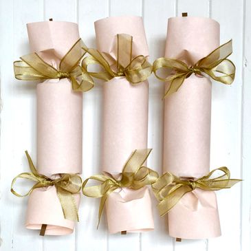 Six pale pink Christmas crackers tied with gold ribbons on a white background.