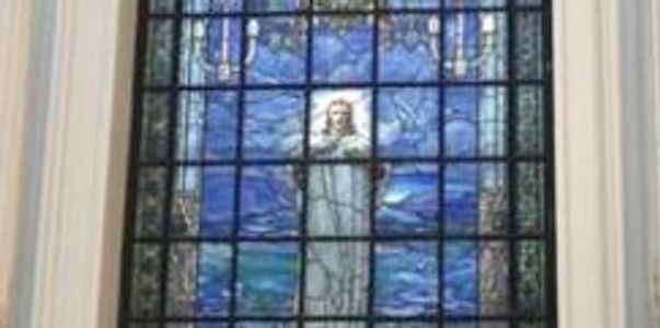 Stained glass window depicting a robed figure with arms raised, surrounded by blue and yellow patterns.