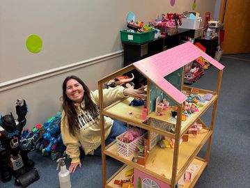 A woman smiles sitting next to a large dollhouse filled with toys.