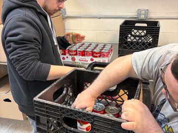 Two men packing canned tomato products into crates in a storage room.