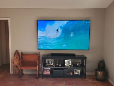 Wall-mounted TV showing a surfer, with a wooden chair and black console below.