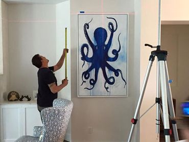 Man measuring a wall with octopus painting and tripod nearby.