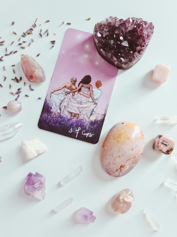 A 3 of Cups tarot card surrounded by various crystals on a white surface.