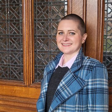 Smiling person with short hair wearing a blue plaid coat standing by a wooden and glass door.