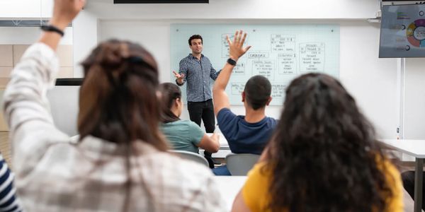 Students raising hands while a teacher explains in a classroom.