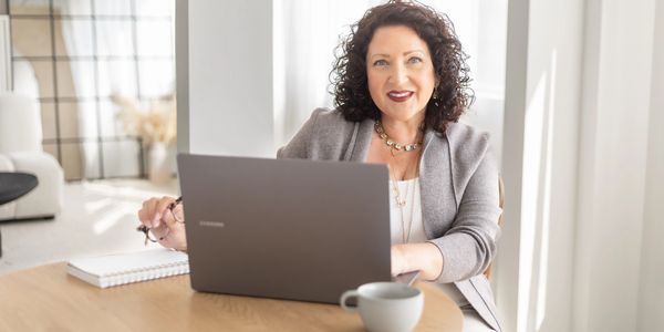 HR Consultant, Maura Richardson, sitting at a desk table with laptop on desk, holding eyeglasses