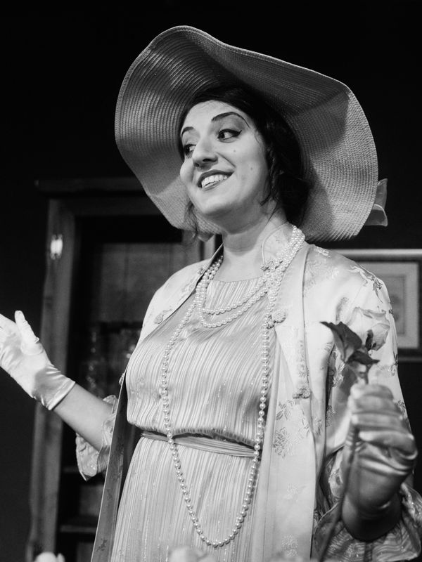 Woman in vintage attire with pearls and hat smiles confidently indoors.