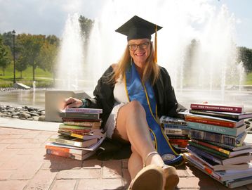 Graduate in cap and gown sits by fountain with stacks of books.