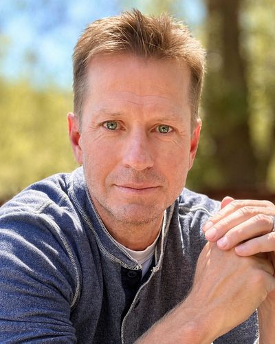 Man with light brown hair and blue eyes outdoors, wearing a gray sweater.