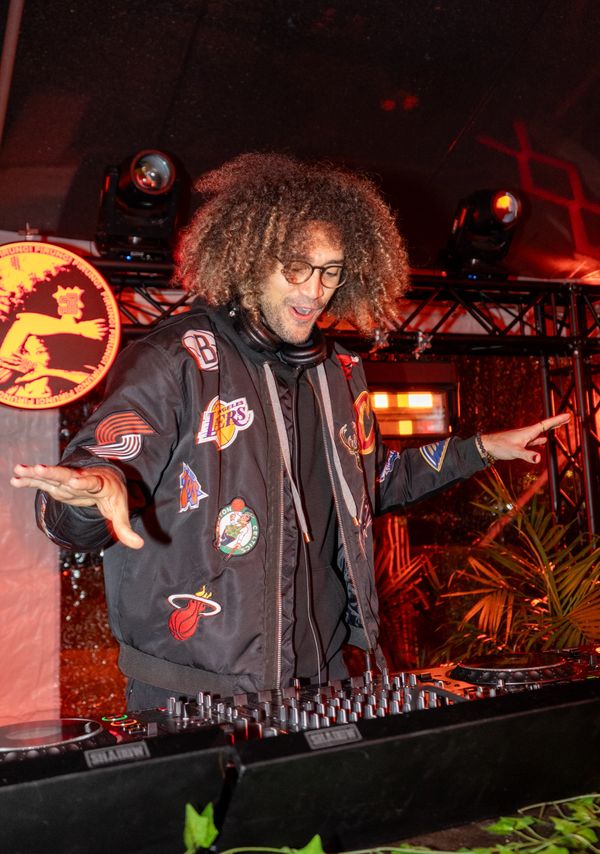 DJ with curly hair and glasses mixing music at a lively event.