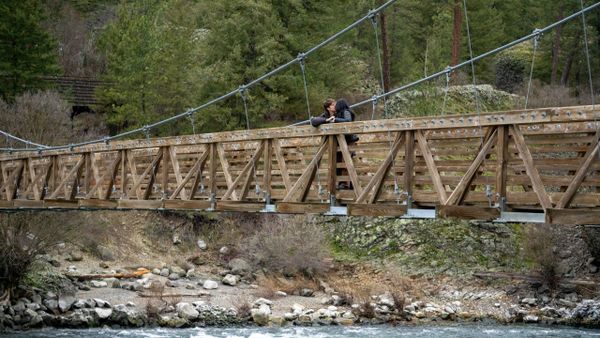 Two people leaning on a wooden bridge railing over a river in a forested area.