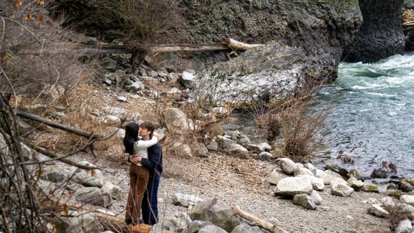 A couple embraces by a rocky riverbank surrounded by nature.